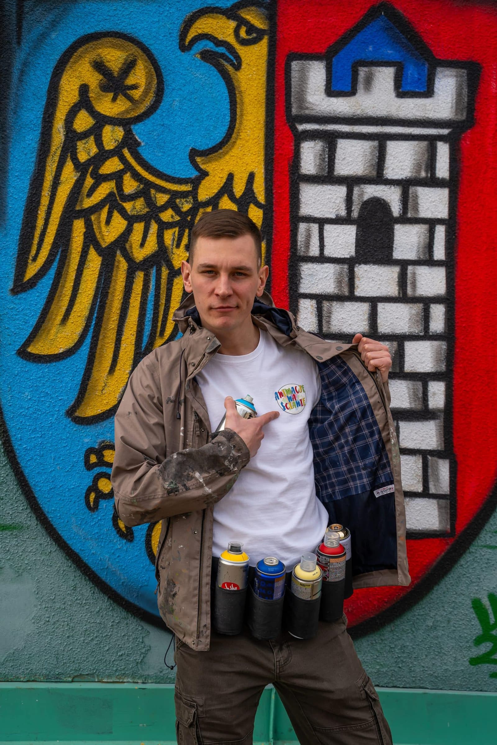 Bartosz Prajsnar - artysta i założyciel Animacje na Ścianie, z farbami w sprayu na tle graffiti z herbem Gliwic