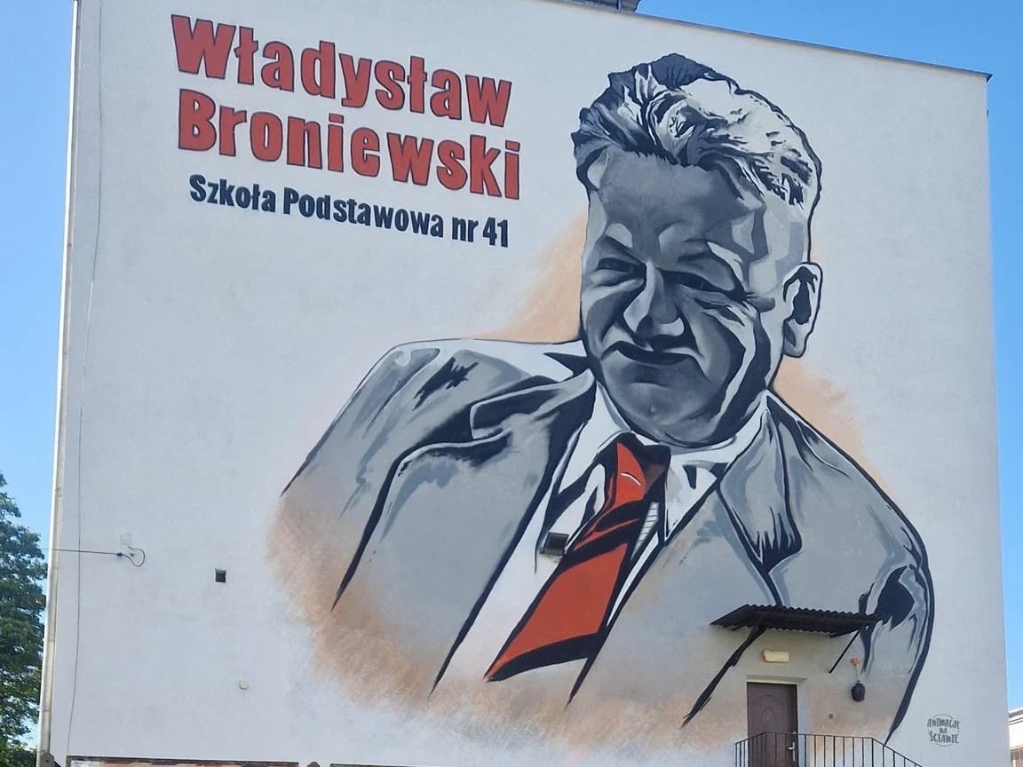 Mural z wizerunkiem patrona szkoły - Władysława Broniewskiego. Utrzymany w odcieniach szarości z pomarańczowymi wstawkami.